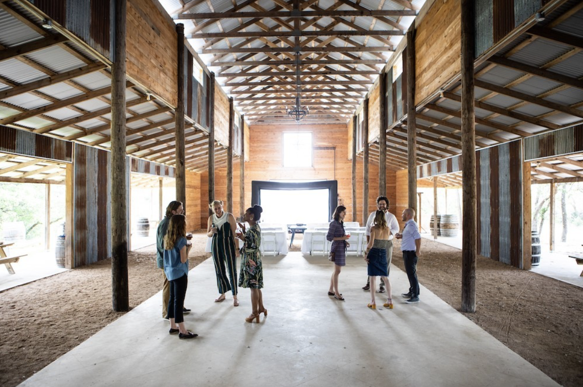 Eight people stand on concrete floor in a large, bright open-air barn.