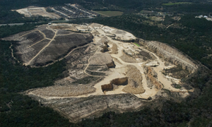 An aerial photograph shows a dramatic view of a massive gravel mine rising out of the woods next to a neighborhood and natural scenery - like a tan-colored scar on the land.