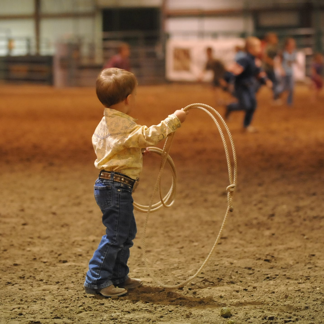 A little boy in cowboy boots and a button down shirt swings a rope in the middle of a rodeo arena.