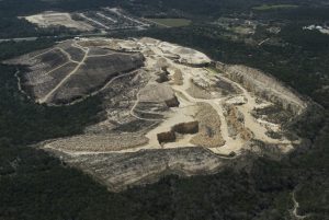 An aerial photograph shows a dramatic view of a massive gravel mine rising out of the woods next to a neighborhood and natural scenery - like a tan-colored scar on the land.