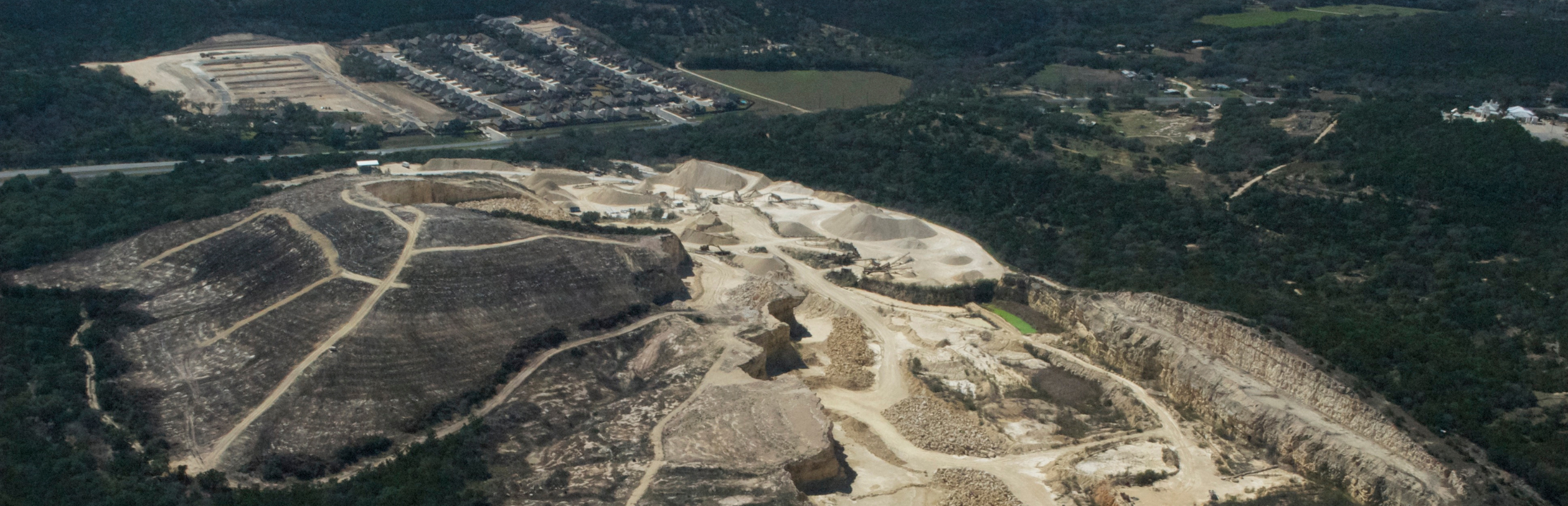 An aerial photograph shows a dramatic view of a massive gravel mine rising out of the woods next to a neighborhood and natural scenery - like a tan-colored scar on the land.