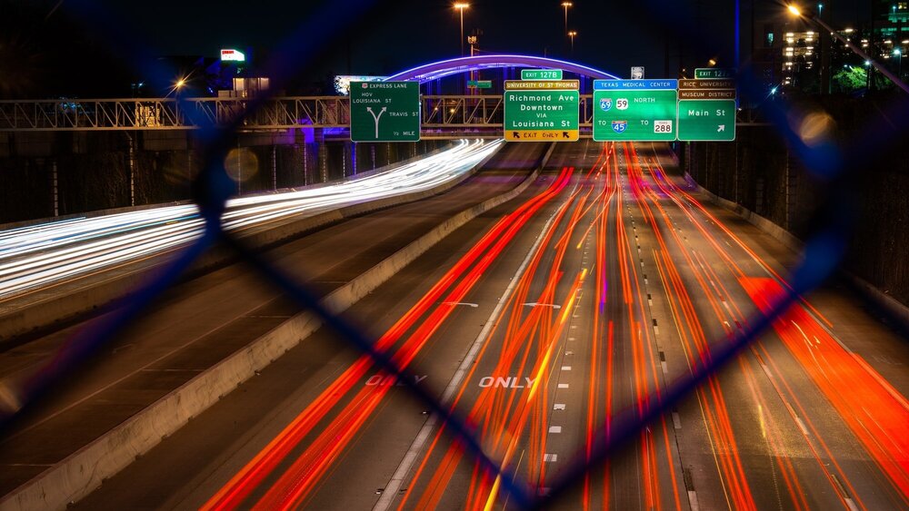 Timelapse photo of a highway at night, with strips of red car lights underneath green exit signs